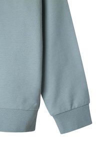 Felpa azzurra chiara con polsini a coste e una texture morbida. Il design presenta una vestibilità rilassata e dettagli di cucitura sottili sulle cuciture.