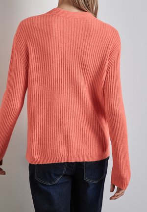 Korallenroter Rippenpullover mit langen Ärmeln, Rundhalsausschnitt und lockerem Schnitt, kombiniert mit dunkelblauen Jeans und einem strukturierten Strickmuster.
