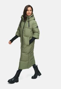 Olivgrön, knälång puffer-jacka med huva, korta ärmar, framlyftning och sidofickor. Kombineras med svarta ankstövlar.