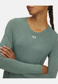 Maglietta da attività a maniche lunghe in verde tenue, in tessuto texturizzato con sottili punte nere, caratterizzata da un colletto rotondo e un logo bianco sul petto.