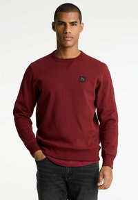 Felpa color bordeaux realizzata in misto cotone, con scollatura a girocollo, polsini a costine e una piccola etichetta nera sul petto.