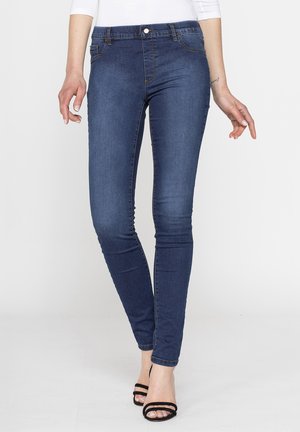 Donna che indossa jeans skinny blu scuro e tacchi neri aperti in punta, in piedi su uno sfondo chiaro e semplice.