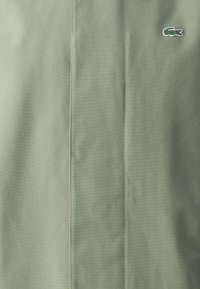 Tessuto verde con una texture liscia e una lucentezza sottile. Presenta una cucitura verticale e un piccolo logo ricamato sul lato sinistro.