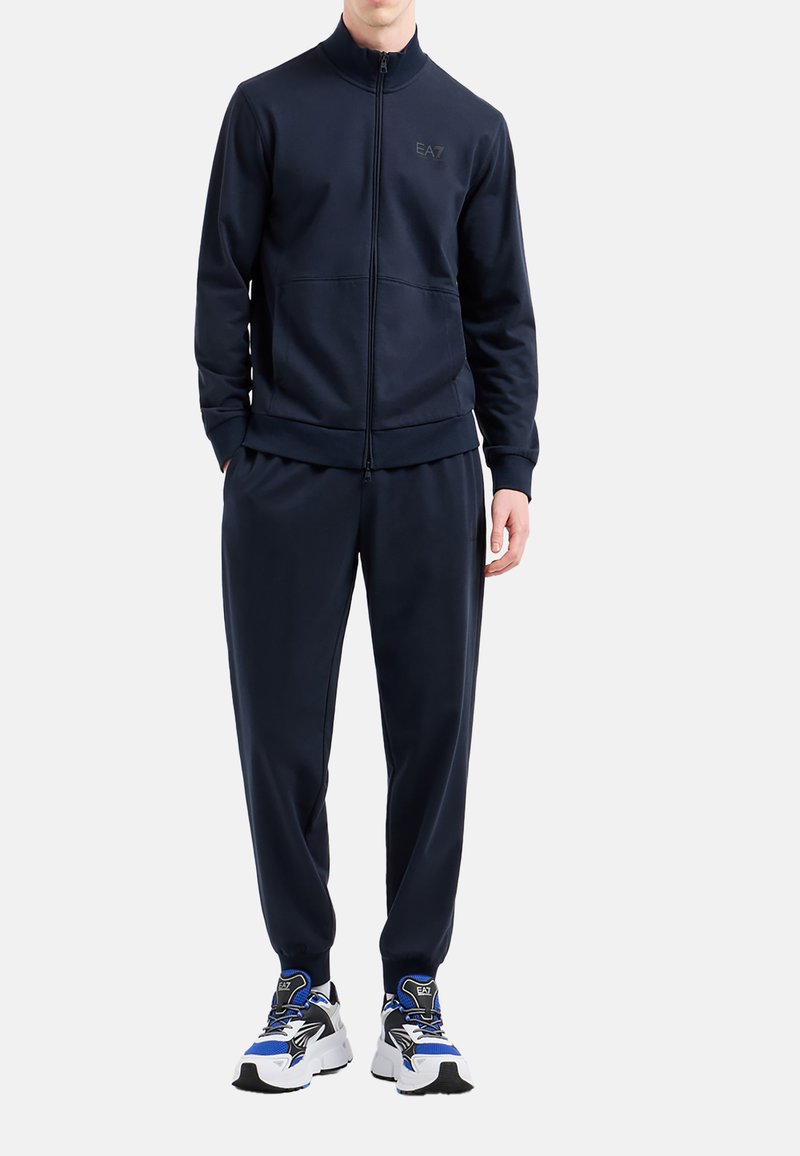 Completo sportivo blu navy con giacca con zip e pantaloni con cintura elastica, caratterizzato da un logo discreto, abbinato a scarpe da ginnastica multicolori.