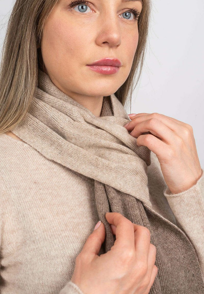 Donna con capelli castani chiari che indossa un maglione beige, mentre sistema una sciarpa bicolore beige e marrone attorno al collo su uno sfondo semplice.