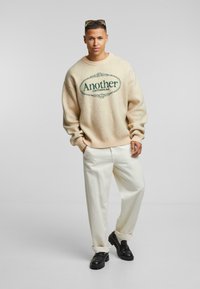 Beżowy sweter dzianinowy z zielonym napisem "Another COTTONLAG", w połączeniu z kremowymi spodniami i czarnymi masywnymi butami. Prosty design przez cały czas.