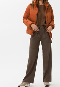 Veste matelassée orange, pull en laine marron, et large pantalon à carreaux marron avec une ceinture. Des baskets beiges avec des accents foncés complètent la tenue.