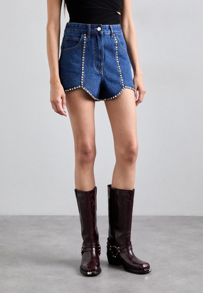 Shorts in denim con orlo scallopato, con dettagli borchiati lungo i lati, abbinati a stivali borgogna alti fino al ginocchio con accenti a fibbia.