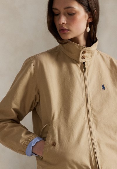 Ung kvinne med lukkede øyne iført beige jakke med glidelås, høy krage og liten blå logo på brystet, hånden i sidelommen.