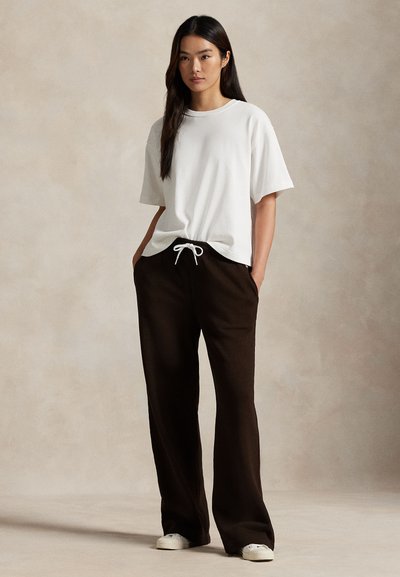 Donna in piedi con le mani nelle tasche, indossa una camicia bianca a maniche corte, pantaloni neri a gamba larga con coulisse e sneakers bianche su sfondo beige.
