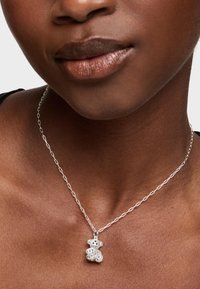 Collier en chaîne argentée avec un pendentif en forme d'ours en peluche, présentant des motifs découpés complexes, reposant sur un teint de peau lisse.