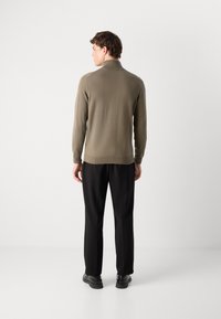 Maglione marrone lavorato a maglia con colletto alto, mostrato dal retro. Abbinato a pantaloni neri e scarpe nere, su uno sfondo semplice.