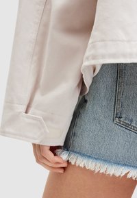 Camisa de algodón de manga larga en un rosa claro y texturizada, con puños doblados y un detalle en parche en la muñeca, combinada con unos pantalones cortos de mezclilla azul claro.