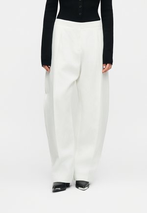 Λευκά παντελόνια με φαρδύ πόδι φορεμένα με μαύρο τοπ με κουμπιά και μαύρα παπούτσια μυτερά με μεταλλικές λεπτομέρειες.