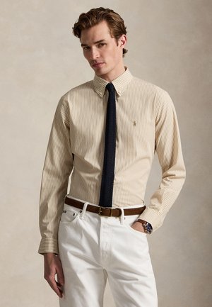Jeune homme portant une chemise à rayures beige, une cravate tricotée bleu marine, un pantalon blanc, une ceinture en cuir marron et une montre-bracelet sur fond neutre.