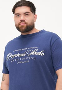 Blue cotton t-shirt featuring a graphic print in white text that reads "Garments Made for Everyday Originals Studio Artist District."