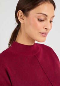 Bordeaux hoge hals trui van zacht gebreid materiaal, met een gestructureerde afwerking en minimalistische naaddetails. Volledige schouderbedekking.