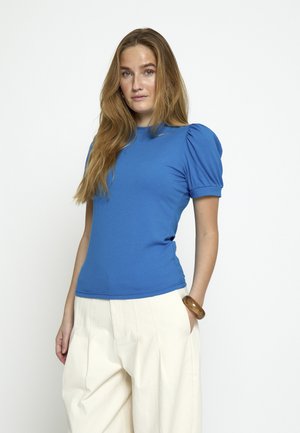 Kobieta o długich blond włosach, ubrana w niebieską bluzkę z krótkim rękawem i białe, szerokie spodnie, stojąca z jedną ręką w kieszeni.