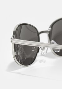 Lunettes de soleil avec une monture en métal argenté, des verres teintés foncés et des coussinets en caoutchouc, au design élégant et minimaliste.