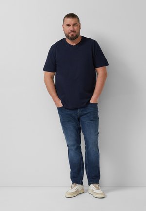 Mann mit Bart und zurückgebundenem Haar steht mit den Händen in den Taschen, trägt ein marineblaues T-Shirt, blaue Jeans und weiße Turnschuhe vor einer schlichten Wand.