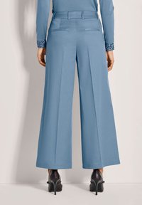 Blaue weit geschnittene Hose mit Falten, flachem Bund und zwei Gesäßtaschen. Getragen mit schwarzen High Heels, die die glatte Stoffoberfläche zur Geltung bringen.