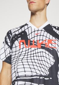 Koszulka sportowa Nike z czarno-białym wzorem w kratę, odważnym czerwonym logo oraz klasycznym dekoltem w serek. Miękki, oddychający materiał.