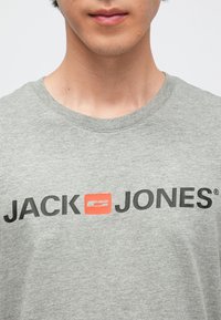 Grå bomulls-T-skjorte med svart "JACK" og "JONES" logo med en oransje aksentboks i midten. Rund hals og korte ermer.