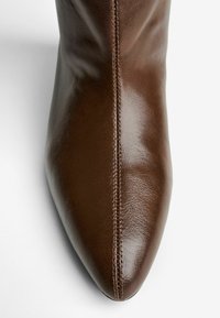 Bottine en cuir marron avec un bout pointu, texture lisse et coutures visibles le long de la couture. Design élégant sans ornementation.