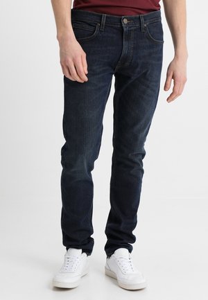 Mies pukeutuneena tummansinisiin slim fit -farkkuihin ja valkoisiin lenkkareihin, seisoo valkoista taustaa vasten.