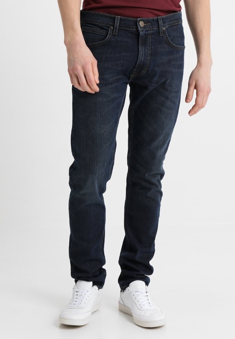 Homme portant un jean skinny bleu foncé et des baskets blanches, debout devant un fond blanc.