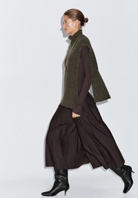 Olive green sleeveless knit top with high neck, paired with a brown flowing skirt and black heeled boots, featuring smooth fabric and minimal design.