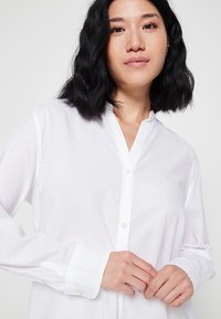Camicia bianca con un design senza colletto, maniche lunghe e polsini con due bottoni. Tessuto liscio, leggero e con un delicato lucido.