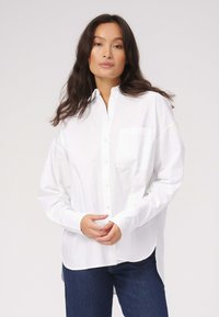 Hvid bomulds skjorte med knapper og lommer; har en afslappet pasform, lange ærmer og buet kant. Bæres med mørkeblå jeans.