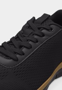 Κοντινή λήψη ενός μαύρου αθλητικού παπουτσιού με υφασμάτινο επάνω μέρος, μαύρα κορδόνια και σόλα σε χρώμα καραμέλας, σε λευκό φόντο.