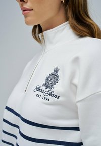 Sudadera blanca con cremallera y rayas horizontales en azul marino, que presenta un logo bordado y el texto "Est. 1994" en un tono azul contrastante.