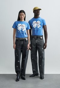 Modré trička s grafikou a kresleným motivem, nošená s černými džínami zdobenými vyšívanými detaily, kombinovaná s černými kotníkovými botami.