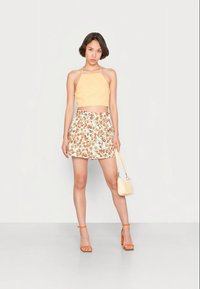 Mulher usando um top halter amarelo, saia branca com estampado de frutas laranja, sandálias de salto alto laranja, segurando uma pequena bolsa bege.