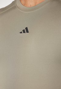 Svijetlo maslinasto zelena atletska majica glatke teksture, kratkih rukava, s crnim logotipom u gornjem lijevom kutu.