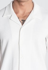 Camisa blanca de textura de gofre con cuello y tres botones delanteros, que presenta un corte relajado y mangas cortas.