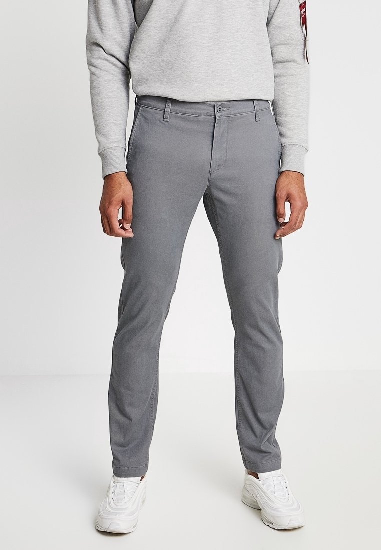 Homme portant un pantalon gris slim, des baskets blanches et un sweat-shirt gris clair, debout devant un fond blanc uni.