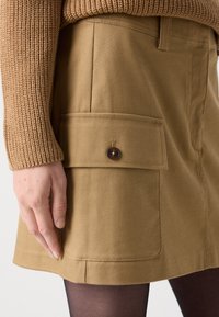 Beige bomulls skjørt med en frontlomme med en brun knapp. Stoffet har et teksturert mønster, og nederkanten er rett.