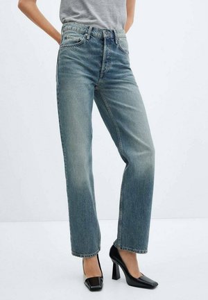 Vrouw die lichtblauwe jeans met rechte pijpen, een grijs shirt en zwarte hoge hakken draagt, staand tegen een witte achtergrond.