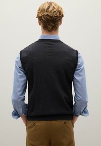 Anthrazitgrauer ärmelloser Pullover über einem hellblauen Langarmhemd, mit glatter Textur und Rundhalsausschnitt, kombiniert mit braunen Hosen.