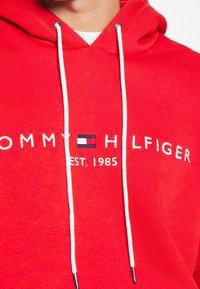 Sudadera roja con cordones, que presenta el texto bordado en blanco "TOMMY HILFIGER EST. 1985" y un pequeño acento del logo en azul marino y rojo.