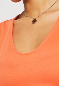 Gros plan d'une personne portant un haut orange à encolure dégagée et un collier chaîne argenté avec un pendentif rond.