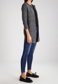 Szary, strukturalny długi kardigan z rękawami trzyczwartowymi, noszony na czarnym topie, w połączeniu z niebieskimi dopasowanymi dżinsami i czarnymi butami z kokardkami.