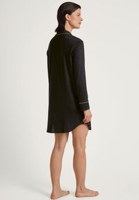 Nightdress nero a maniche lunghe realizzato in tessuto morbido. Presenta un colletto, piping dorato sui polsini e un orlo curvo.