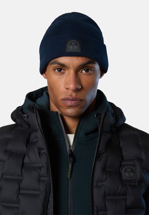 Uomo con berretto navy e giacca trapuntata nera con logo North Sails visibile su berretto e giacca, espressione facciale neutra.