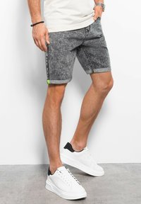 Jeansshorts in zwart en grijs met een opgerolde zoom, voorzien van een klein groen accentlabel. Gecombineerd met witte sneakers met een zwarte hiel.