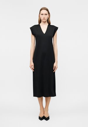 Γυναίκα που φοράει μαύρο midi φόρεμα χωρίς μανίκια με λαιμόκοψη σε σχήμα V, στέκεται όρθια μπροστά σε απλό ανοιχτό γκρι φόντο.